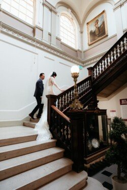 A bride and groom ascend a grand staircase with ornate railings inside the high-ceilinged Guildhall Bath, featuring large windows and a distinguished portrait on the wall.