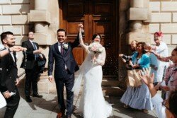 A bride and groom walk out of the stunning Guildhall in Bath holding hands, greeted by guests who throw confetti. The groom wears a dark suit, and the bride is in a white dress holding a bouquet.