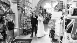 A bride and groom walk through an indoor market filled with various merchandise, including a stand with postcards and a shop displaying textiles, reminiscent of the charming atmosphere found at Guildhall Bath Weddings.