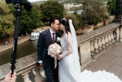 A couple dressed in wedding attire shares an intimate moment on a stone balcony at Guildhall Bath Weddings, overlooking a river with green trees in the background. A photographer's equipment is partially visible on the left side, capturing their special day.