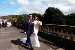 A bride and groom walk hand in hand on a sunlit stone pathway at Guildhall Bath Weddings, surrounded by lush greenery and a partly cloudy sky. The bride is in a white dress, holding a bouquet; the groom wears a dark suit.