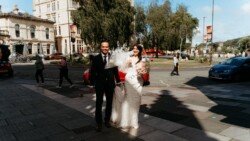 A bride in a white wedding dress and a groom in a dark suit walk together outdoors on a sunny day, with the historic charm of Guildhall Bath Weddings and trees in the background.