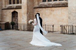 A bride and groom walk outside the historic Guildhall Bath, with the bride in a white gown and long veil, holding a bouquet of flowers.