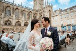 A newlywed couple in wedding attire stands arm in arm, smiling at each other in front of a historic cathedral with people sitting on benches in the background, capturing the essence of Guildhall Bath Weddings.
