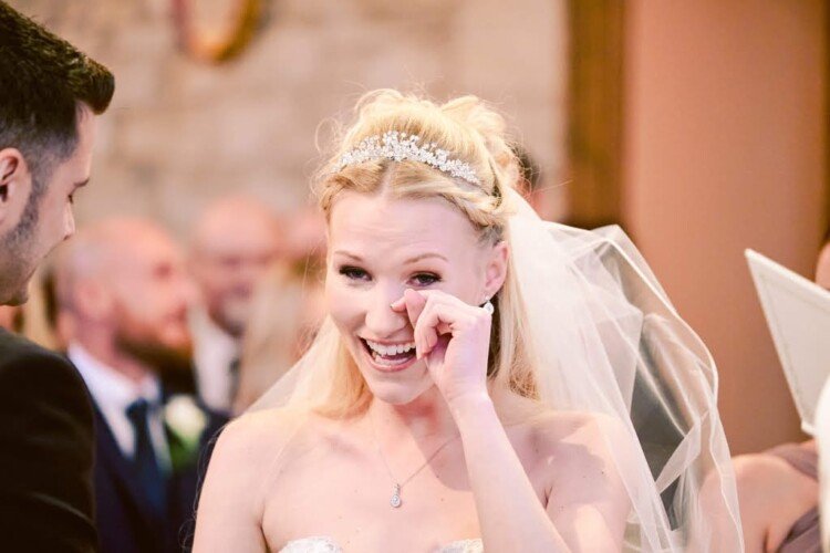 A bride in a white dress and tiara wipes a tear from her eye while smiling during the wedding ceremony, captured beautifully by the photographer in Bradford on Avon. The groom and guests stand in the background, sharing this heartfelt moment.