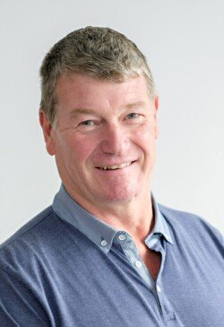 A middle-aged man with short gray hair is smiling while wearing a blue polo shirt with a light blue collar, perfect for corporate photography. He stands against a plain light background.