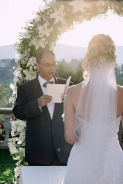 A man in a suit reads from a booklet under a floral arch at an Italian wedding, while a bride in a white dress with a veil stands facing him outdoors.