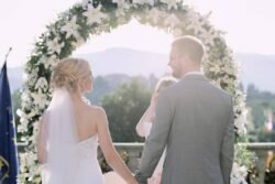 A bride and groom stand hand in hand in front of a flower arch during an outdoor Italian wedding ceremony, facing each other and smiling.