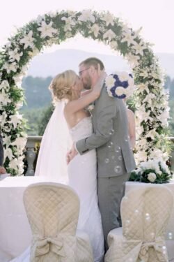 A bride and groom share a kiss in front of a floral arch at an outdoor Italian wedding ceremony, with decorative chairs and bubbles floating in the foreground.