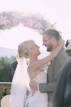 A bride and groom embrace and smile at each other under a floral arch during a romantic Italian wedding ceremony outdoors.