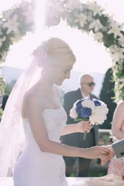 A bride in a white gown and veil holds a bouquet of blue and white flowers during an outdoor Italian wedding ceremony, with sunlight streaming through a floral arch.