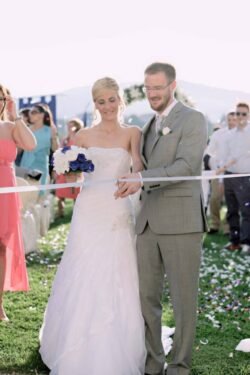 A bride and groom stand together outdoors, cutting a white ribbon with scissors, surrounded by guests at their beautiful Italian wedding ceremony.