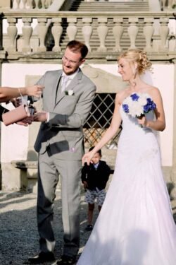 At an Italian wedding, a bride in a white dress and a groom in a gray suit stand outdoors, receiving a small item from a guest as a child walks behind them.