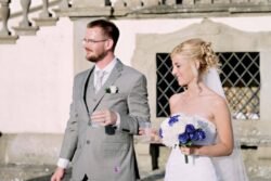 At an Italian wedding, a bride in a strapless white dress holds a bouquet of white and purple flowers; her groom in a light grey suit stands beside her. Both are holding drinks outdoors.