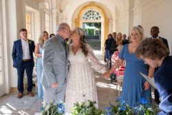 A couple is kissing indoors during their wedding ceremony at Hestercombe House & Gardens, with guests standing and seated around them. The bride holds a bouquet, and the groom is dressed in a grey suit. The setting is well-lit with natural light.