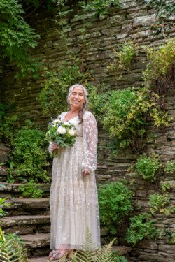 A woman in a light, floral-patterned dress stands on stone steps at Hestercombe House & Gardens, holding a bouquet of flowers, with a backdrop of lush greenery and a stone wall.