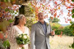 A couple stands outdoors under the fall foliage at Hestercombe House & Gardens, smiling at each other. The woman holds a bouquet of white roses while the man holds a glass of champagne. Both are dressed in formal attire, adding to the elegance of their picturesque surroundings.