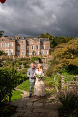 A couple walks down a stone path in Hestercombe House & Gardens, with an old mansion and stormy sky in the background. The woman holds a bouquet and wears a long dress; the man wears a light-colored suit.