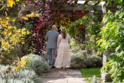 A couple walking under a wooden arbor adorned with red and green foliage at Hestercombe House & Gardens, surrounded by lush garden scenery. The individual on the right is wearing a white dress, and the one on the left is in a gray suit.