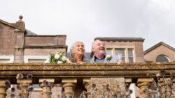 An elderly couple stands on a stone balcony, smiling and holding a bouquet and glass, with the historic Hestercombe House & Gardens in the background.