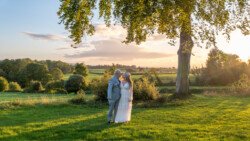 A couple stands closely together in a lush green field at sunset, with trees and the distant landscape of Hestercombe House & Gardens in the background.