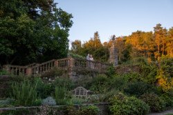 A couple stands on a stone terrace at Hestercombe House & Gardens, overlooking a lush garden with various plants and trees during the golden hour.