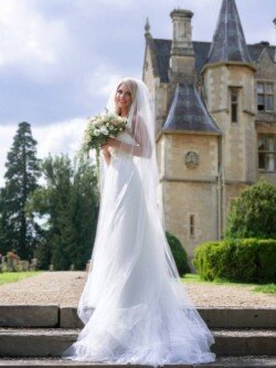 OLYMPUS DIGITAL CAMERA A bride dressed in a white wedding gown and veil holds a bouquet of flowers while standing in front of the historic, castle-like building at Orchardleigh Weddings on a sunny day.