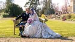 A bride in a white gown and a groom in a kilt and jacket sit on a wooden bench in the garden at Orchardleigh Weddings, smiling and holding a bouquet.
