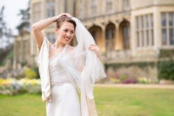A bride in a white wedding gown and veil stands outdoors in front of a large, ornate building at Orchardleigh Weddings, smiling and adjusting her veil. The setting includes green grass and blooming flowers.