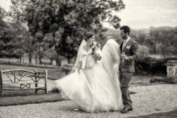 A bride and groom walk together on a gravel path outdoors at Orchardleigh Weddings, with the bride holding her gown and a bouquet, the groom holding a drink. Both are smiling. There is a wooden bench and trees in the background.