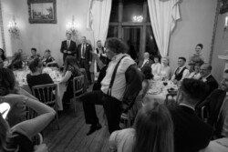 At an Orchardleigh Weddings event, a man stands and performs a playful dance in the middle of a formal gathering, surrounded by seated guests dressed in elegant attire, in an exquisitely decorated room.