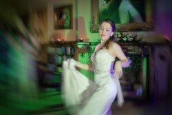 A woman in a white dress is captured spinning gracefully indoors, her eyes closed, and holding part of her dress. The background shows a blurred room with bookshelves and framed pictures, evoking the enchanting ambiance typical of Orchardleigh Weddings.