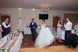 A bride and groom walk into a reception hall as guests stand and applaud. The bride is in a white gown, the groom in a blue suit. The beautifully decorated hall exudes the charm reminiscent of Orchardleigh Weddings.