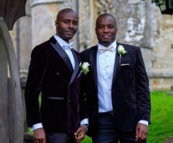 Two men in formal attire with white bow ties and boutonnières stand outdoors by a stone building, exuding the elegance typical of Orchardleigh Weddings.