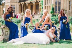A bride in a white dress reclines on the grass, surrounded by six bridesmaids in royal blue dresses holding bouquets, posing in front of a historic building at Orchardleigh Weddings.
