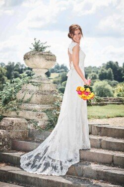 A woman in a white wedding dress stands on stone steps outdoors, holding a bouquet of colorful flowers and smiling at the camera. Behind her, the lush greenery and cloudy sky create a serene backdrop typical of Orchardleigh Weddings.