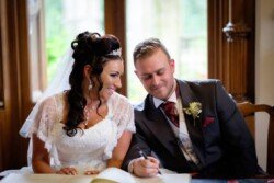 A bride in a white dress and a groom in a dark suit smile as the groom signs a document at an Orchardleigh Weddings event indoors.
