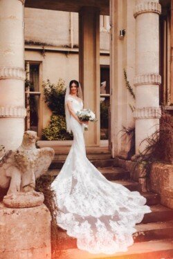 A bride in a white gown with a long lace train stands on steps in front of a stone building, holding a bouquet of flowers and smiling, capturing the essence of Orchardleigh Weddings.