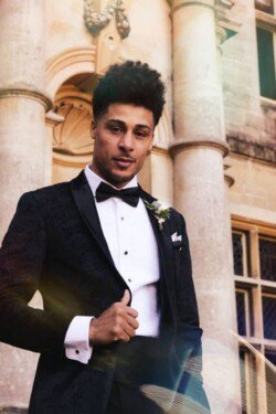 A man in a black tuxedo with a white shirt and black bow tie stands in front of a stone building with columns, holding his jacket lapel. A small white boutonniere is pinned to his lapel, capturing the timeless elegance that defines Orchardleigh Weddings.