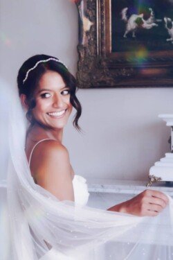 A woman in a wedding dress and veil smiles, standing next to a white mantel with a framed painting above it at her beautiful Orchardleigh Weddings venue.