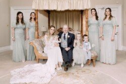 A wedding party poses indoors at Orchardleigh Weddings. The bride and groom sit on a couch with a baby and two young children, while five bridesmaids in matching dresses stand behind them.