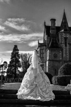 www.thefxworks.co.uk A person wearing a wedding dress and veil stands on stone steps with their back to the camera, facing a gothic-style building and trees in the background under a partly cloudy sky, capturing the enchanting essence of Orchardleigh Weddings.