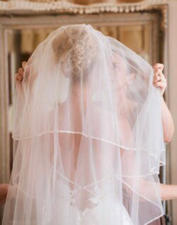 A bride with curly blonde hair is having a white veil placed over her head, assisted by another person, visible from behind. The scene captures the enchanting charm typical of Orchardleigh Weddings.