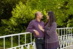 A couple stands on a white metal bridge surrounded by lush greenery, smiling at each other. Engaging in portrait photography, the man wears a purple shirt, and the woman in a patterned dress rests her hand on his chest.