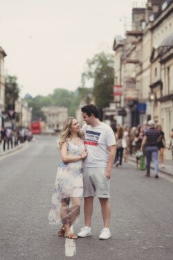 A couple stands in the middle of a street lined with buildings, looking at each other. The woman is wearing a floral dress, and the man is in a white t-shirt and grey shorts. People walk in the background, creating a perfect moment for portrait photography.