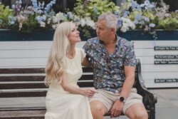 A woman in a light dress and a man in a patterned shirt and shorts sit on a bench, engaged in conversation. Flowers provide a picturesque background, capturing a perfect moment for portrait photography.