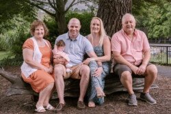 Five people, including one holding a baby, are seated on a large wooden bench outdoors. Capturing the essence of portrait photography, trees and greenery provide a serene backdrop.