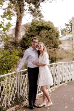 A couple stands together on a pathway beside a white fence, embraced with greenery and a building in the background. The woman wears a white dress and the man wears a light blue shirt and dark pants—a perfect example of portrait photography.