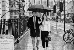 A man and a woman stroll together under an umbrella on a rainy street, passing "The Bath Humbug Shop" sign. The man is holding coffee, and they both appear to be smiling. This charming moment captures the essence of portrait photography, highlighting their joy amidst the drizzle.