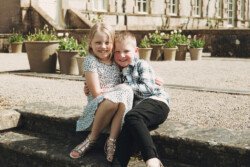 Two children, a boy and a girl, sitting closely together on stone steps, smiling. The background features potted plants and a large building, creating a charming scene of portrait photography.